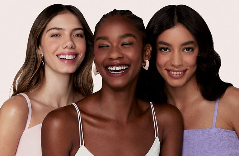 Foto de tres mujeres con diferentes tipos de cabello. El fondo es beige claro, y llevan ropa en tonos pastel: rosa, blanco y lila.